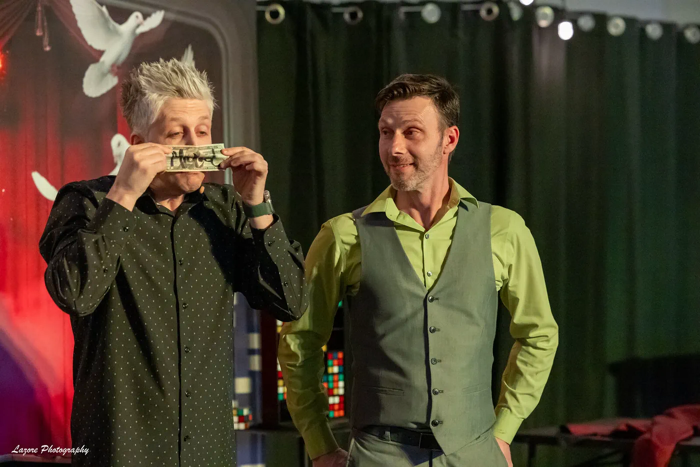 Wade Live on stage playing a borrowed signed bill like a harmonica while a grinning volunteer looks on
