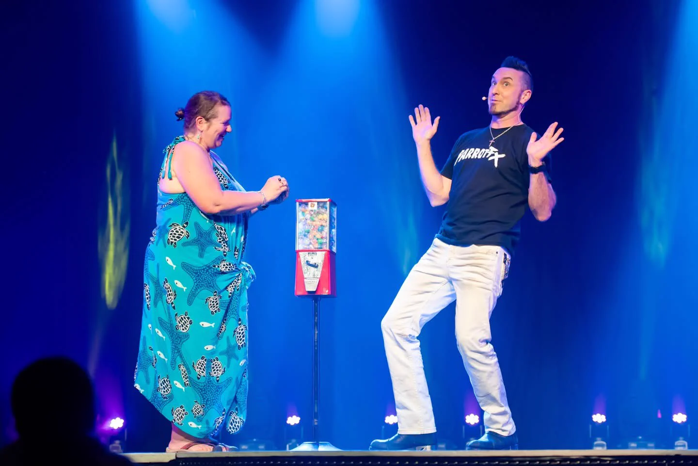 David DaVinci on a blue lit stage, hands up in mock surprise beside The Machine while a volunteer holds what she has just taken out of it
